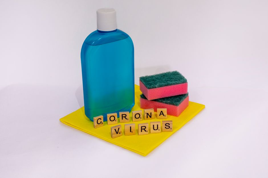 A blue plastic bottle of cleaning solution and three pink and green scrubbing sponges are placed on a yellow cloth on a white surface. In front of the items, yellow Scrabble tiles spell out 'CORONA VI